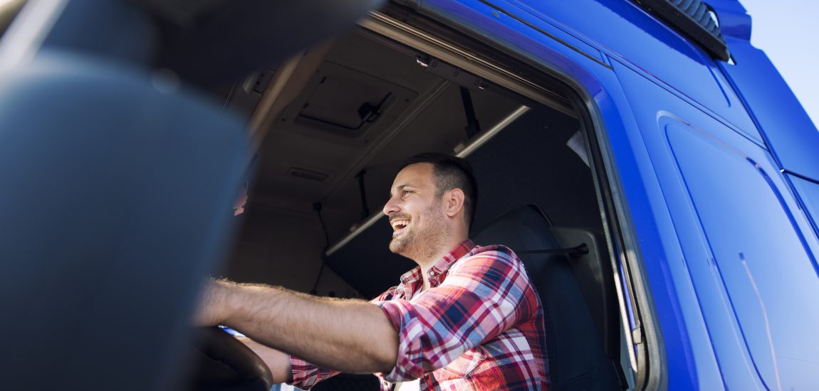 Professional middle aged trucker in cabin driving truck and smiling.